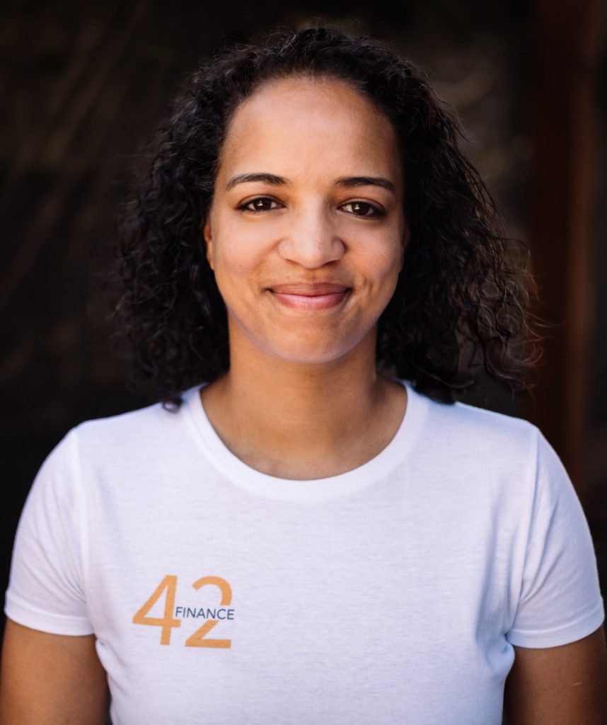 Porträt von Kim, die in die Kamera lächelt. Kim hat dunkle Locken und trägt ein weißes Tshirt mit dem 42Finance Logo